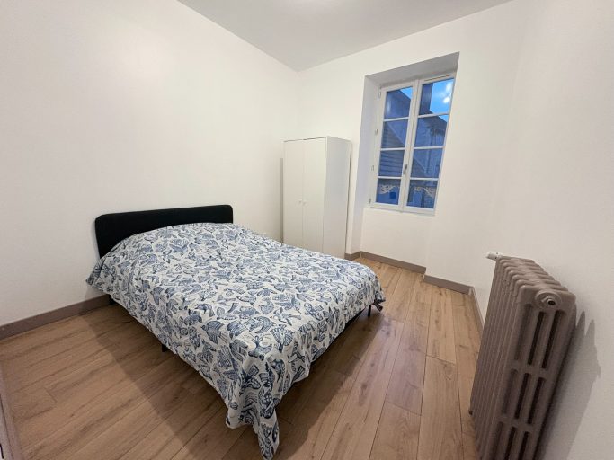 Chambre simple avec un lit, une fenêtre et un radiateur, mur blanc et sol en bois.